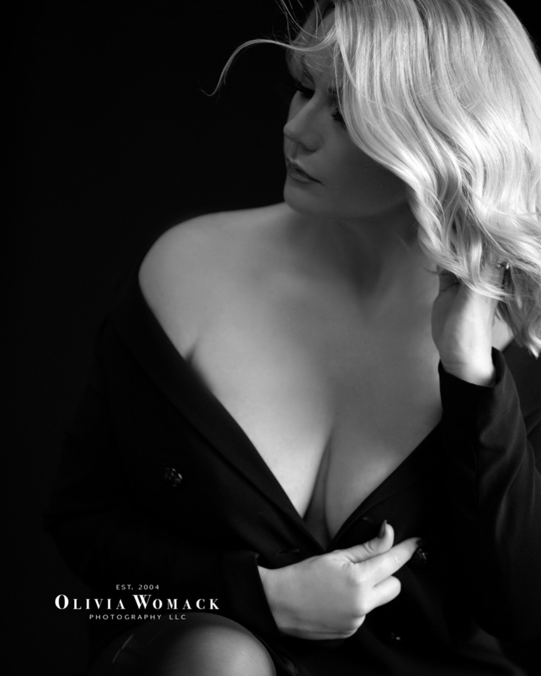 Black and white portrait of a woman with blonde hair, wearing a dark jacket off her shoulders, gazing to her left, hand touching her hair. logo of olivia womack photography llc is visible. Image by Olivia Womack Photography - Merritt Island, FL.
