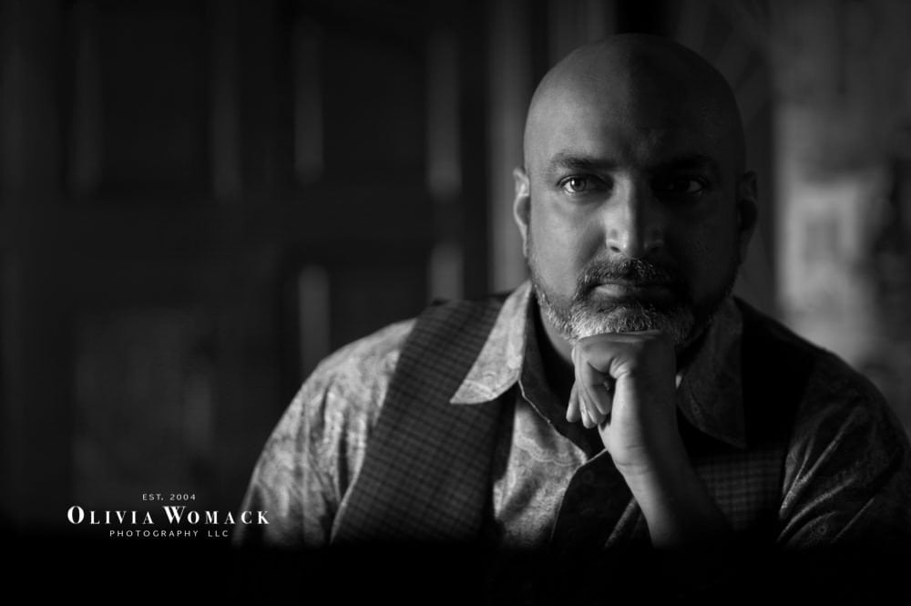 A black and white portrait of a bald man with a beard, resting his chin on his hand, looking intensely towards the camera. the background is blurred and dark, focusing attention on his expression. Image by Olivia Womack Photography - Merritt Island, FL.