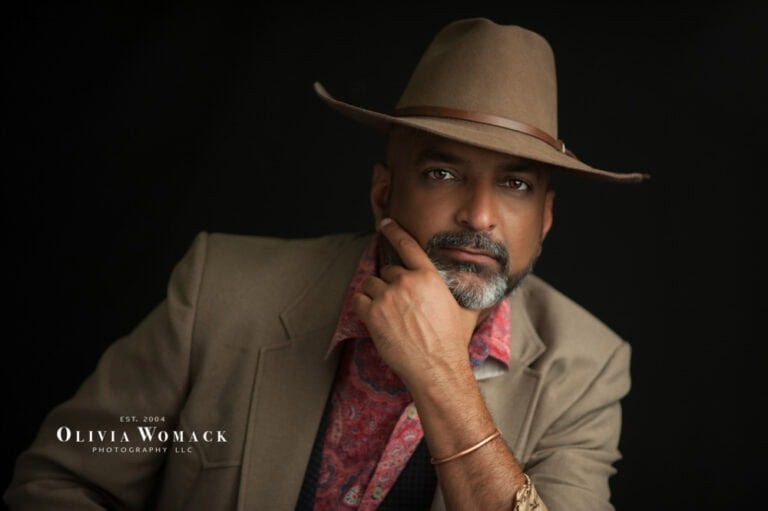 A portrait of a man with a beard wearing a hat, gazing thoughtfully at the camera, with his hand on his chin. he has a scarf around his neck. . Image by Olivia Womack Photography - Merritt Island, FL.
