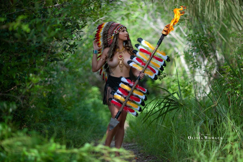 A woman in tribal attire, holding a colorful feathered staff and a flaming torch, stands in a lush green forest pathway. by Olivia Womack Photography