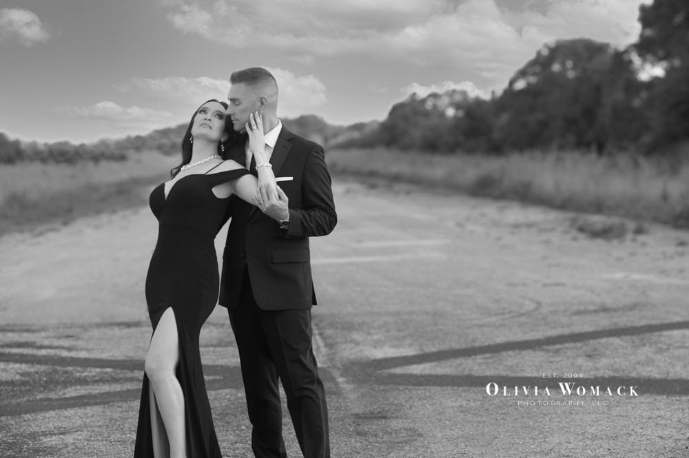 A black-and-white photo of an elegant couple embracing on an empty road with a dramatic sky in the background. the woman in a black gown leans into a man in a suit holding an origami crane. by Olivia Womack Photography