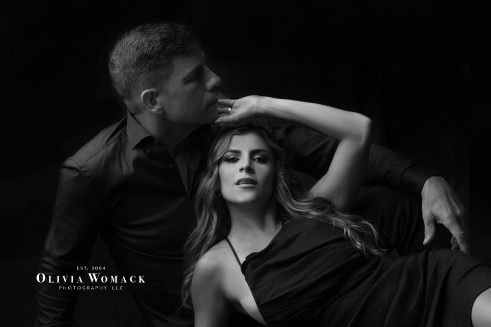 A black and white artistic portrait of a man and a woman in a dramatic pose, with the man standing and the woman reclining, both dressed in elegant dark attire. by Olivia Womack Photography