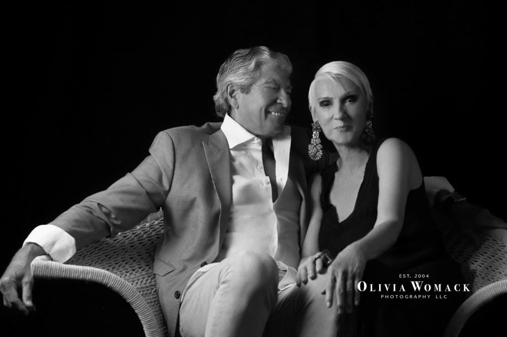 Black and white image of an older couple smiling and sitting closely on a wicker bench, dressed in elegant attire, against a dark backdrop. the man is in a suit, and the woman has an ornate earring. by Olivia Womack Photography