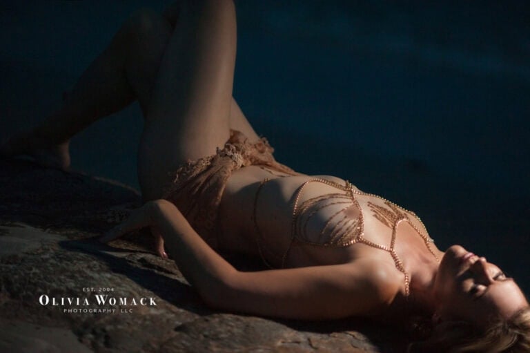 A woman in delicate lingerie lies on a rocky surface, illuminated by soft, dim light highlighting her relaxed pose and contemplative expression. by Olivia Womack Photography