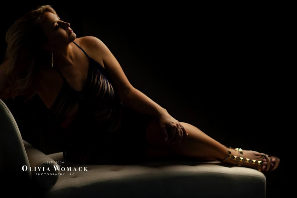 A woman reclines on a couch, illuminated by soft light, wearing a dark dress and strappy sandals, evoking a mood of contemplation and elegance. her blonde hair cascades gently down her shoulders