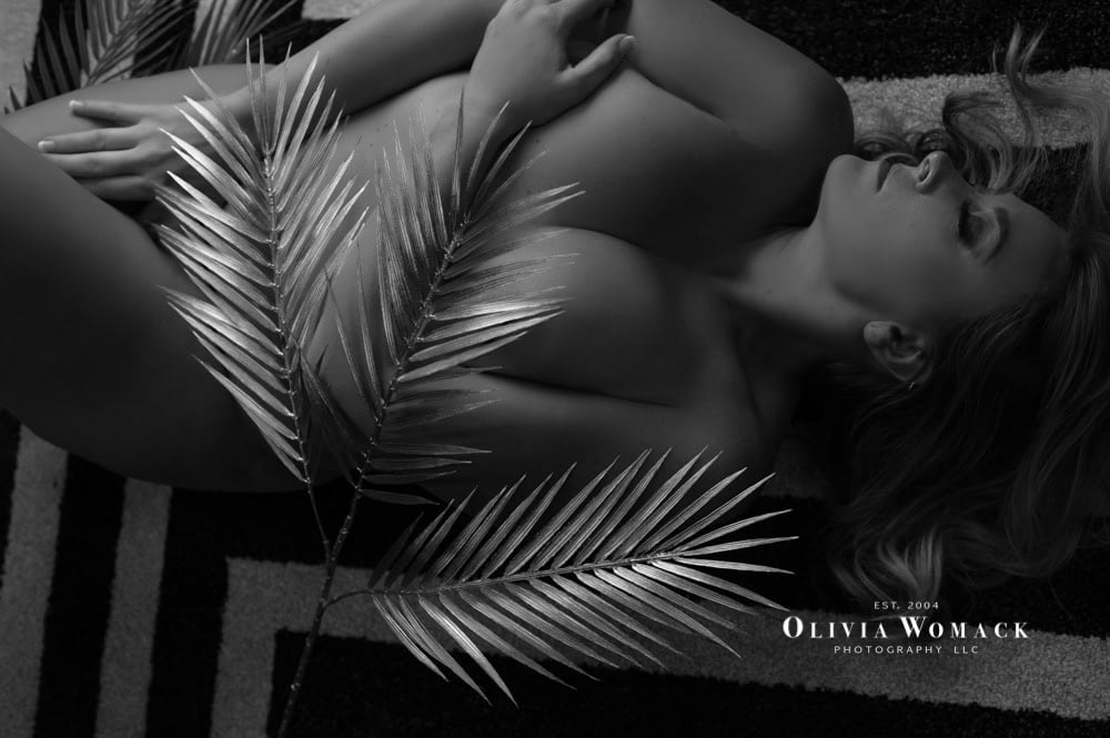 Black and white artistic image of a woman lying on a striped rug, covered with large palm leaves, conveying a serene and elegant mood. photographer's watermark included. by Olivia Womack Photography
